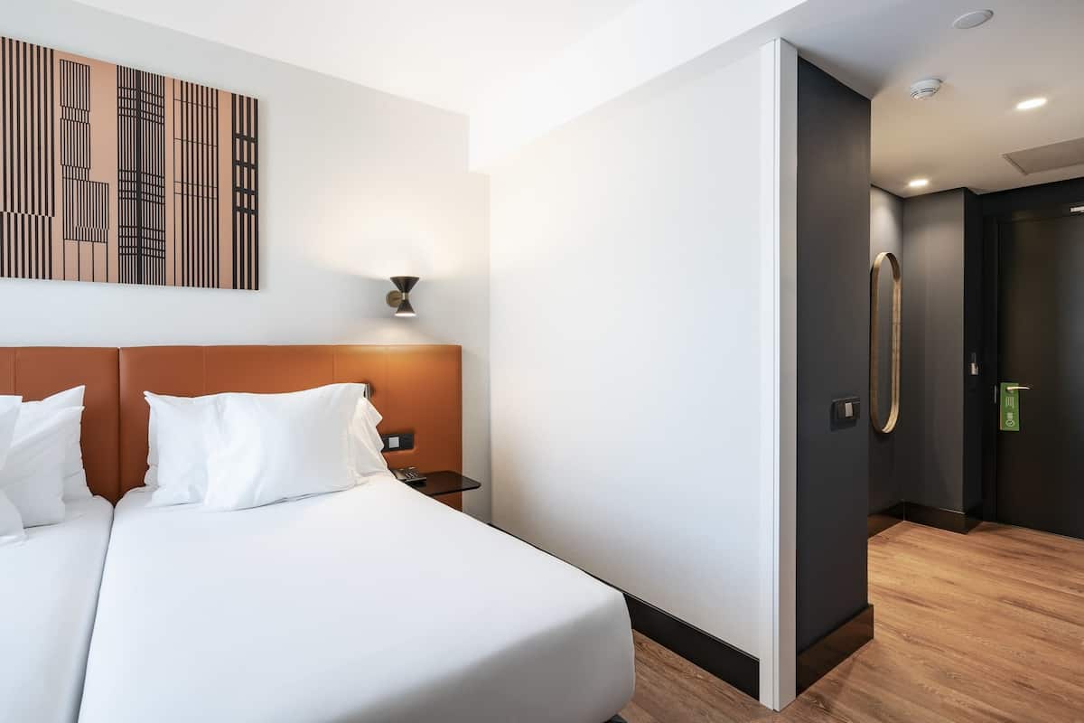 Una habitación de hotel moderna con una cama grande, cabecero, una obra de arte colgada en la pared y una puerta que da al baño.