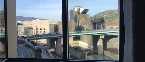 Vista a través de una ventana que muestra un paisaje urbano con un edificio moderno, un puente y una zona peatonal.