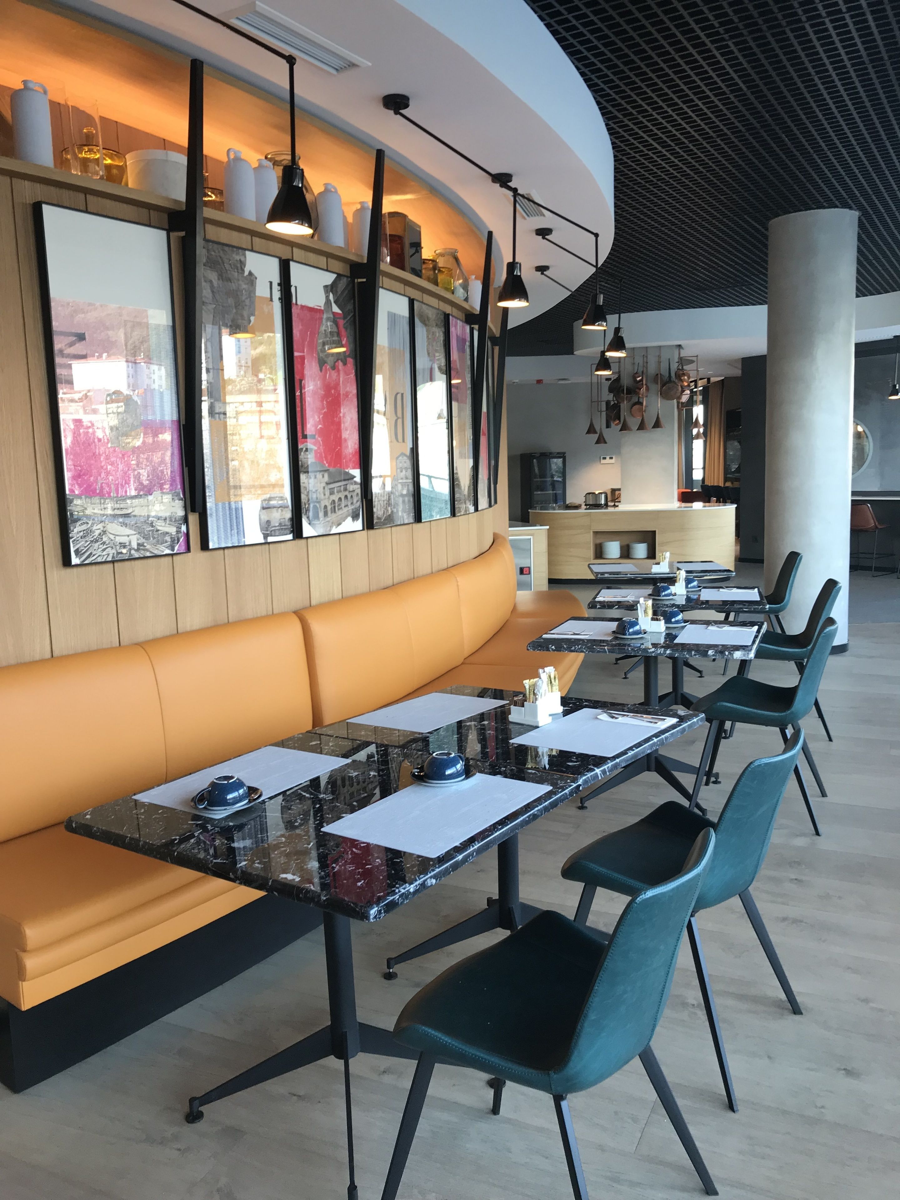 Un restaurante moderno con un área de asientos curva, mesas de mármol negro, sillas color turquesa y una cocina al fondo.