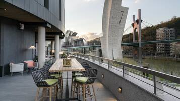 An outdoor seating area with a long wooden table and chairs, overlooking a river and a bridge.