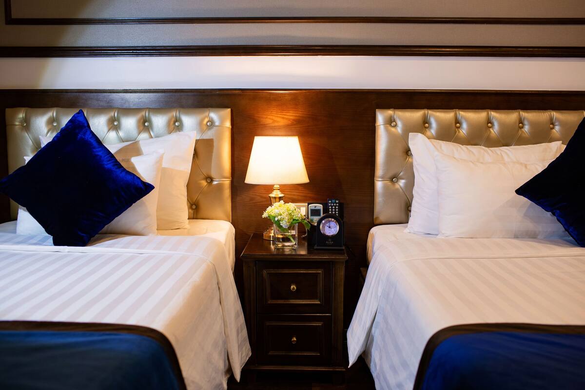 A hotel room with two beds, each with a headboard, a bedside table with a lamp, a clock, and a vase with flowers.