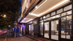 Exterior de un hotel de noche con señalización iluminada, entrada visible y zona de estar con candelabro.