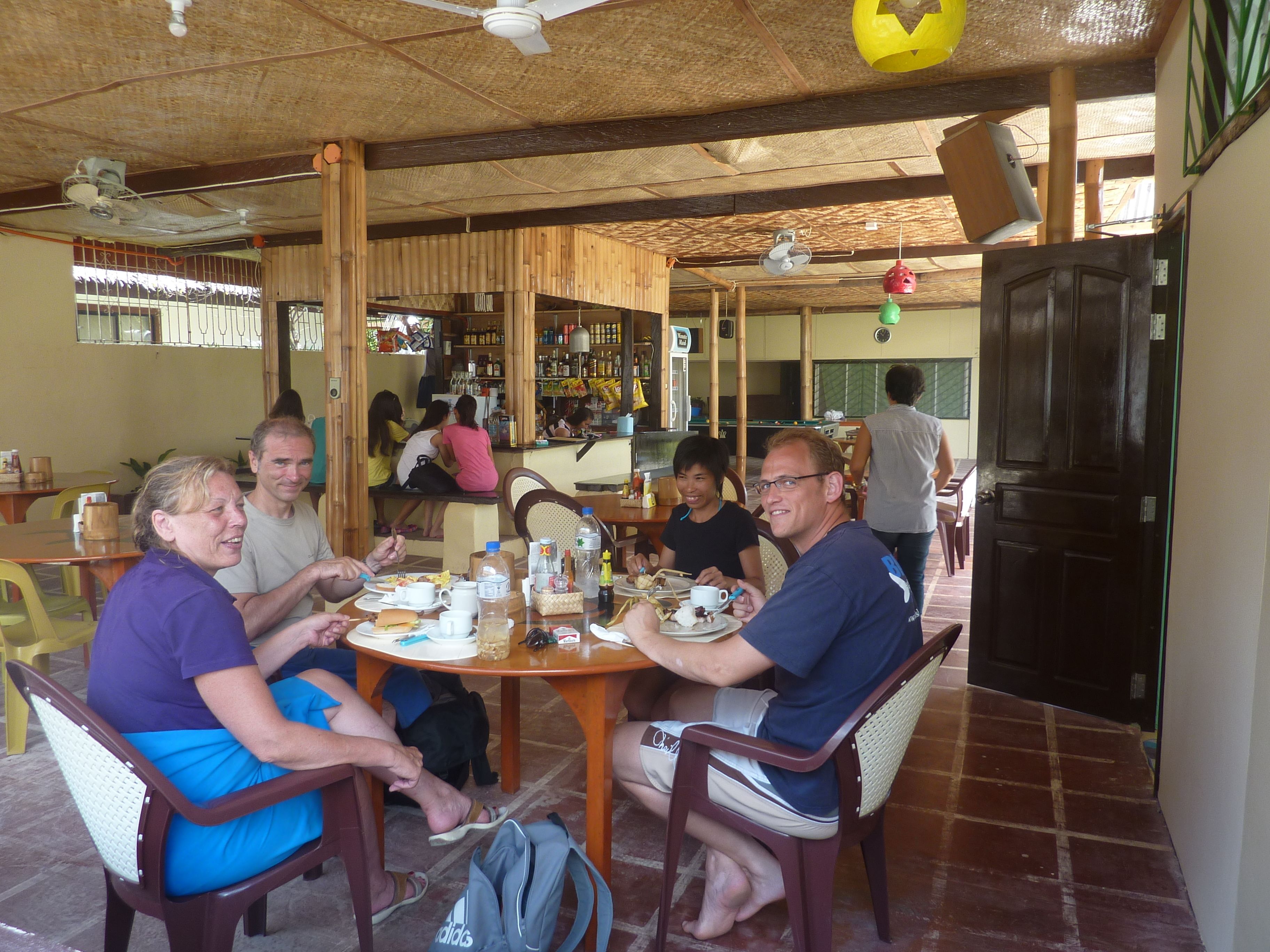 A group of people dining at a wooden table in a rustic setting with a thatched roof and wooden beams.