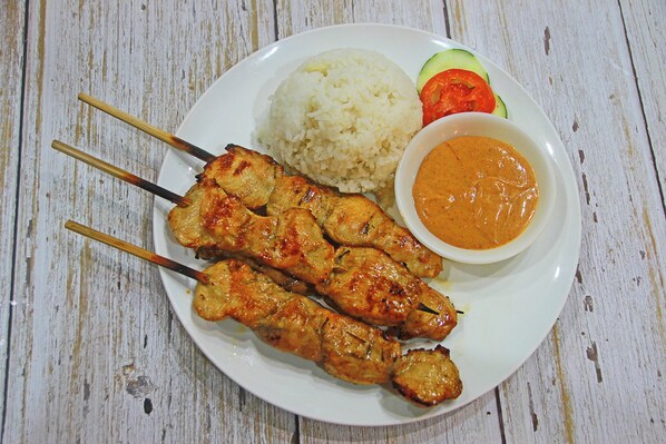 A plate with grilled chicken skewers, white rice, sliced cucumbers, tomatoes, and a bowl of sauce. - Blue Abyss Resort (Moalboal)