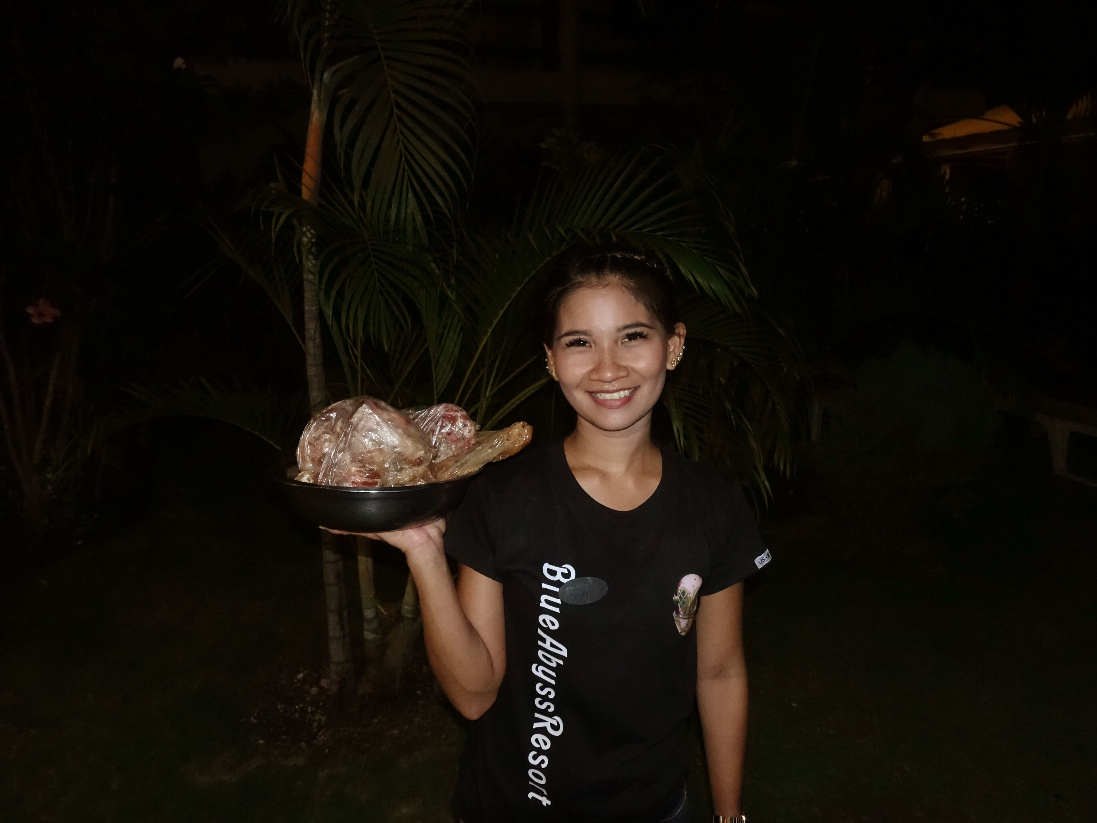 A person holding a bowl with food, wearing a black t-shirt with text, outdoors at night with palm trees in the background.