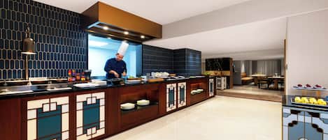 A modern kitchen with a chef preparing food, a dining area with a table and chairs, and a buffet with various dishes.
