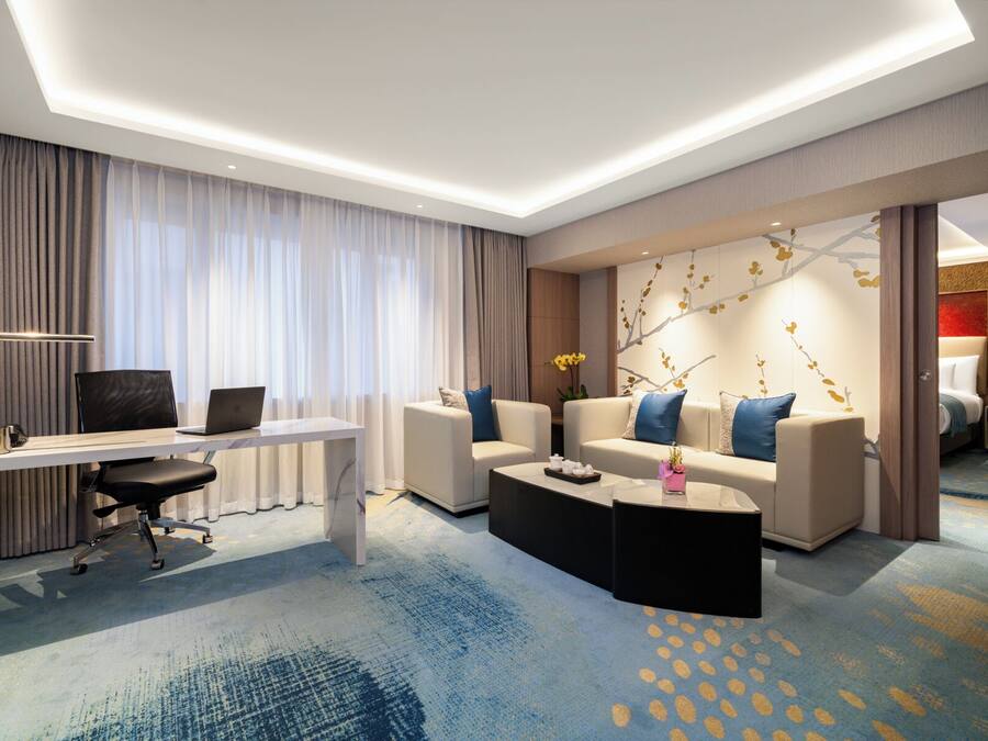 A modern hotel room with a desk, two sofas, a coffee table, and a large window with curtains.