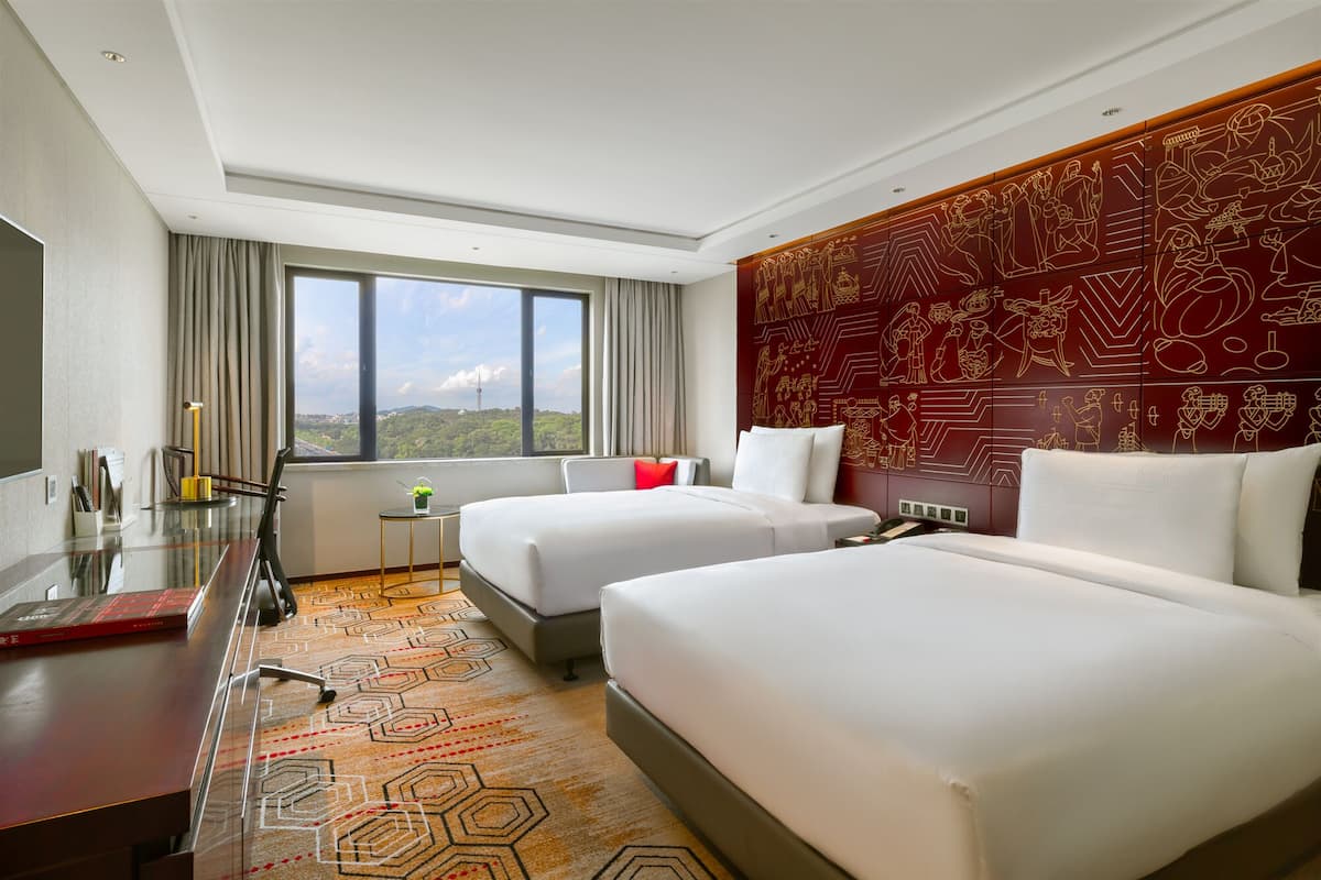 Ein Hotelzimmer mit zwei Betten, einem Schreibtisch, einem Stuhl, einem kleinen Tisch und einem großen Fenster mit Blick auf die Stadt.