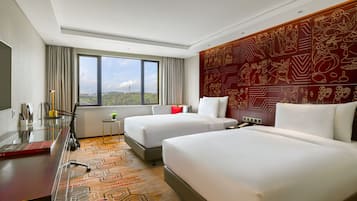 A hotel room with two beds, a desk, a chair, a small table, and a large window offering a city view.