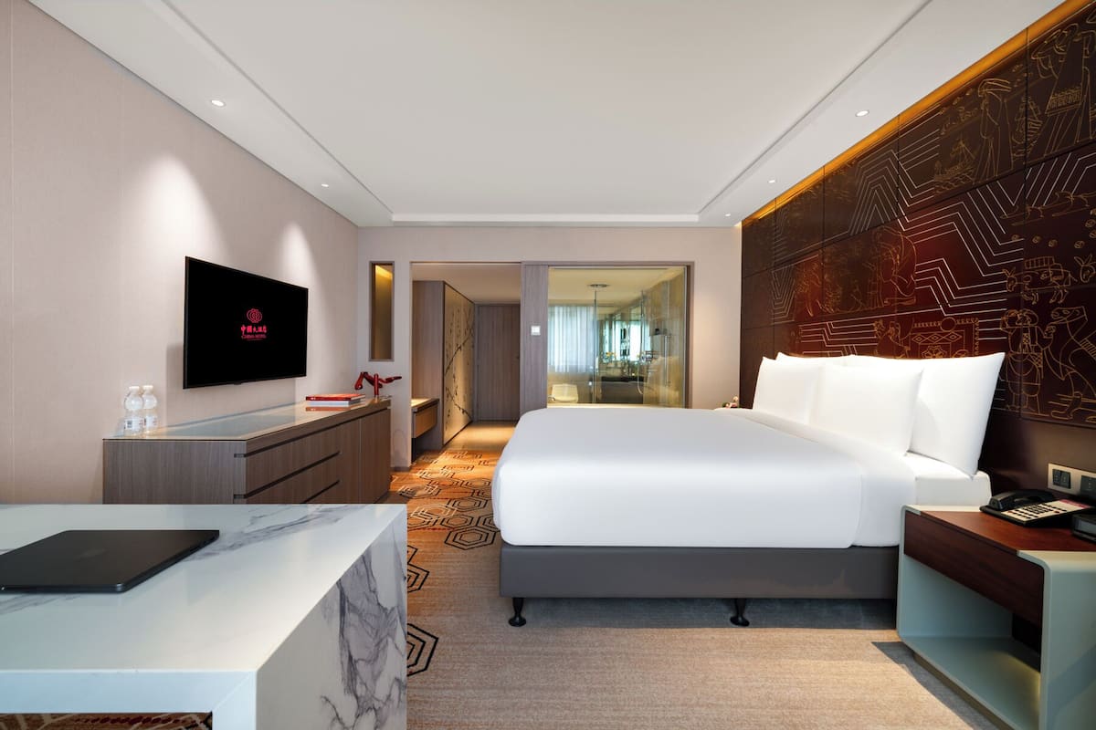 Ein modernes Hotelzimmer mit einem großen Bett, einem Schreibtisch mit einem Laptop, einem Fernseher und einem durch eine Glastür einsehbarer Badezimmer.