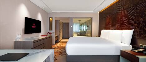 Quarto de hotel moderno com uma cama grande, uma escrivaninha com um laptop, uma televisão e um banheiro visível através de uma porta de vidro.
