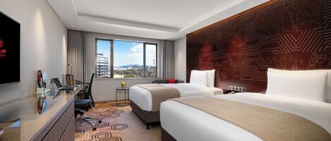 A hotel room with two beds, a desk, a TV, and a view of the cityscape.