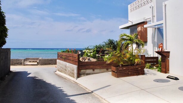 A coastal property with a paved driveway leading to a wooden gate, a white building with a balcony, and a view of the ocean. - Shirahama no yado shiokaze (Nakijin)