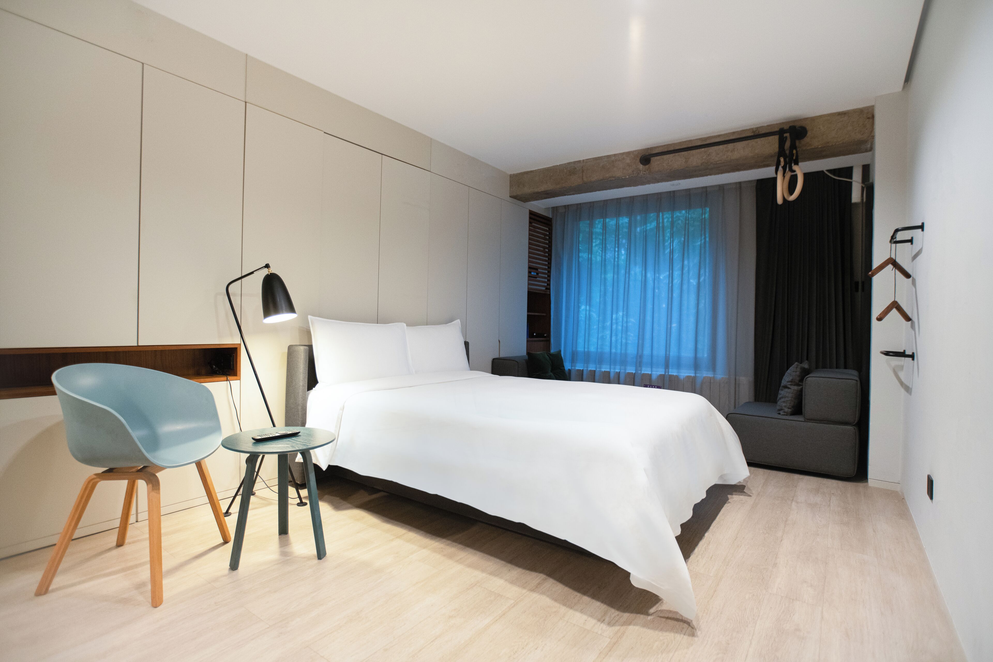 A modern hotel room with a large bed, a desk with a chair, a standing lamp, and a view of the outdoors through a window with curtains.