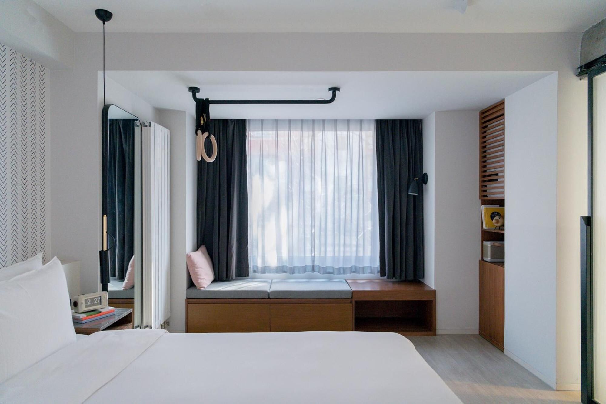 A modern hotel room with a large bed, a window seat with curtains, a desk with a lamp, and a wardrobe.