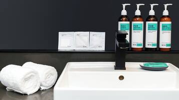 Un baño con un lavabo blanco, un grifo negro y cuatro botellas de productos de cuidado corporal Metis. También hay dos toallas blancas enrolladas y un pequeño plato con un objeto verde.