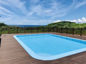 A rectangular swimming pool with a wooden deck and a wooden fence, surrounded by lush greenery and a clear sky. - Private villa LUXE (Nakijin)