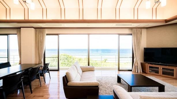 A spacious living room with a large sofa, a dining table, and a flat-screen TV. - Private villa LUXE (Nakijin)
