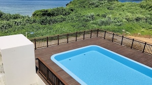 A rectangular swimming pool with a wooden deck and a wooden railing. The pool is surrounded by lush greenery and overlooks a body of water. - Private villa LUXE (Nakijin)