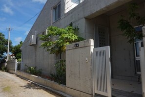 Un bâtiment en béton avec une entrée fermée par un portail, un petit auvent et un arbre qui pousse près de l’entrée.