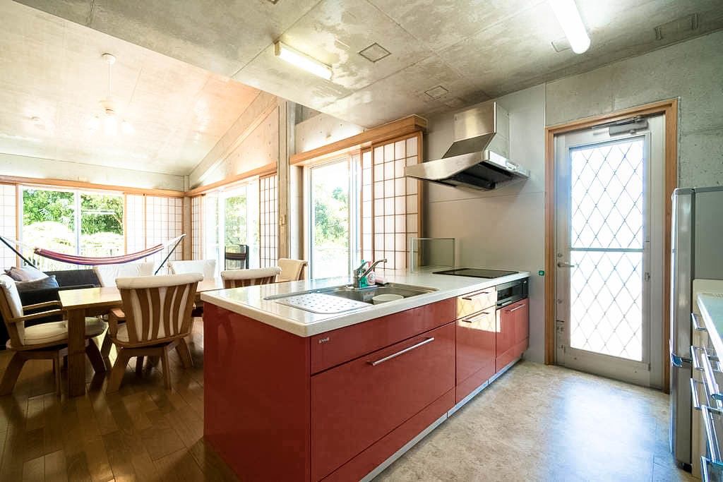 Une cuisine avec un îlot central rouge, des chaises en bois, une table à manger et de grandes fenêtres.