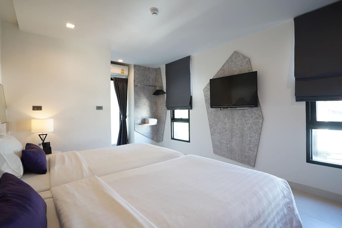 A modern hotel room with two beds, a flat-screen TV mounted on the wall, a window with a view, and a ceiling-mounted air conditioning unit.