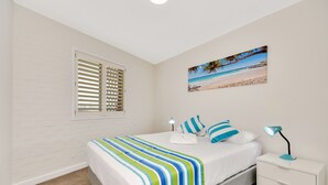 Un dormitorio con cama, mesita de noche, lámpara, ventana con persianas y un cuadro con temática playera en la pared.