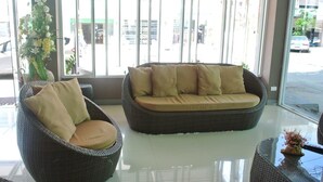 A wicker sofa with cushions, a wicker chair, and a vase with flowers in a lobby area with large windows. - JJ Villa (Khon Kaen)