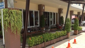 A hotel entrance with a covered seating area, a planter with greenery, and a row of orange traffic cones. - JJ Villa (Khon Kaen)