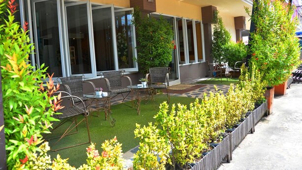 An outdoor seating area with wicker furniture and a small table, surrounded by potted plants and a neatly trimmed lawn. - JJ Villa (Khon Kaen)