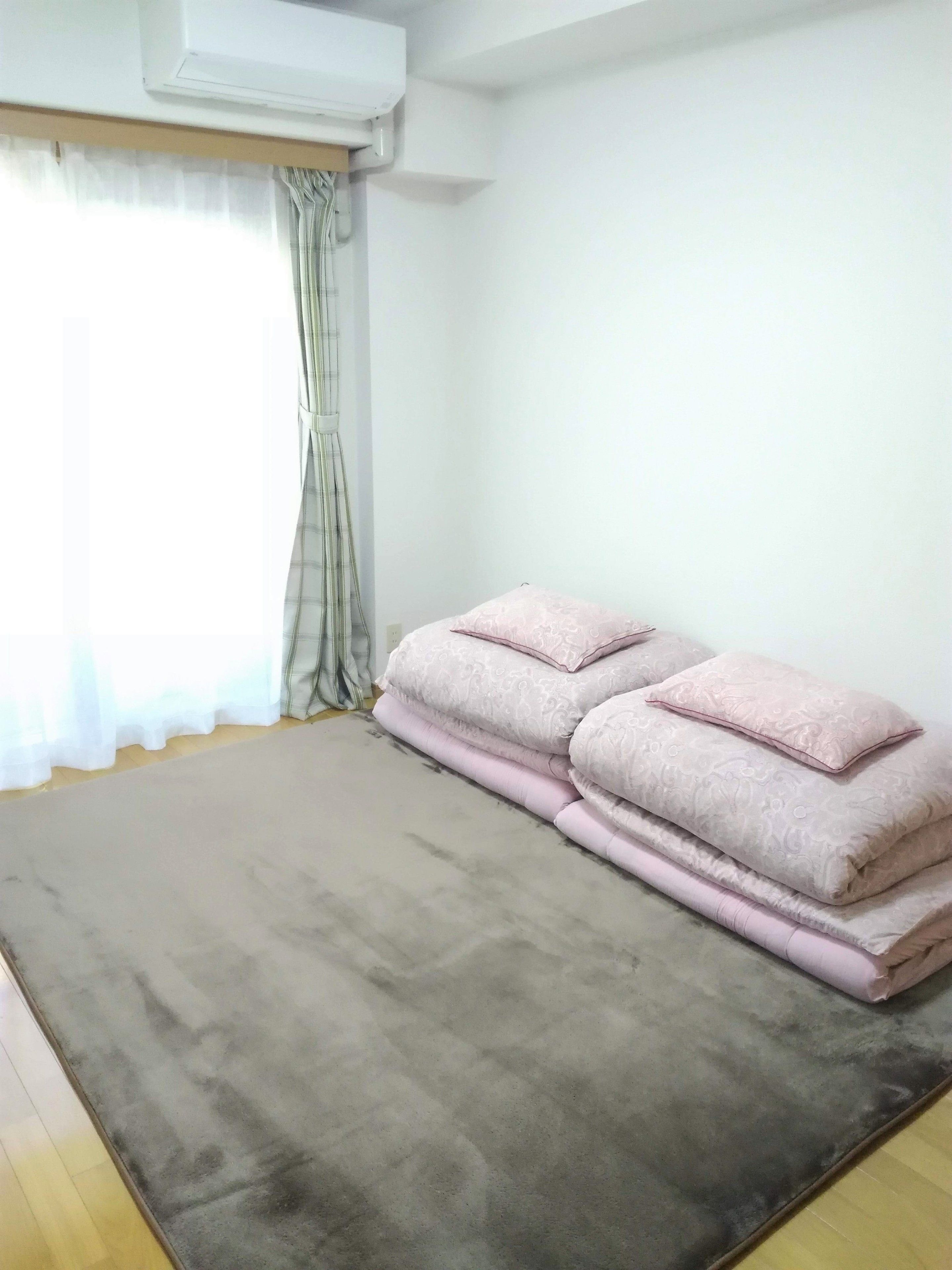 A room with a large grey carpet, a window with sheer curtains, and a stack of pink pillows.