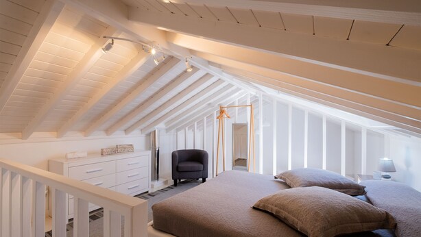 A bedroom with a sloped ceiling, a bed with a brown bedspread, a chair, a nightstand with a lamp, and a dresser. - Casa Vista da Marquesa, Açores - Ideal house for a couple to enjoy nature! (Nordeste)