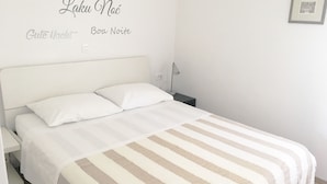 Una cama bien hecha con una colcha a rayas, una mesita de noche y una pared con letreros multilingües que dicen “Buenas noches”.