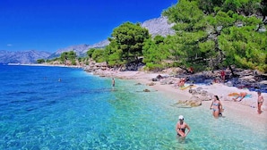 Una playa con agua cristalina, gente nadando y tomando el sol, y árboles en la orilla.