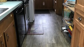 A compact kitchen with a refrigerator, stove, and sink. There's a wooden cabinet with a mirror and a shelf with dishes. A window with curtains is visible, and a rug with a vintage design is on the floor. - Melody's Bed and "Catch Your Breakfast!" (Crockett)