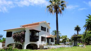 Un edificio blanco con techo de tejas, un balcón con barandilla de madera oscura y una palmera al fondo.