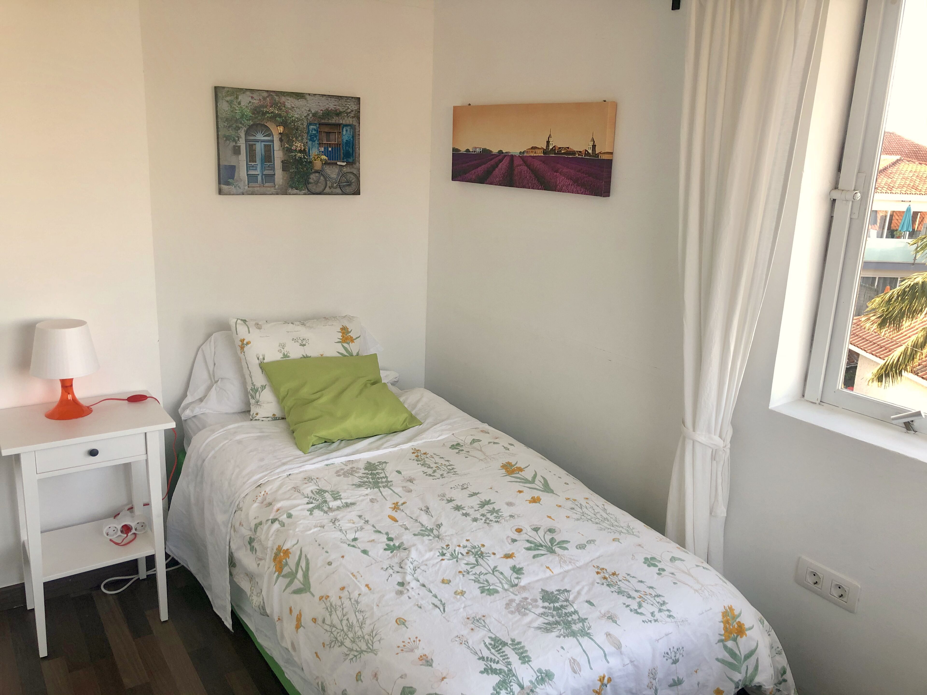 Una camera da letto piccola e ordinata con un letto singolo, un comodino con una lampada e due quadri appesi alle pareti.