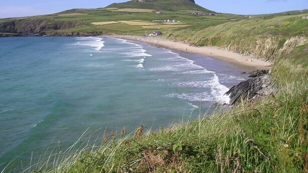 A coastal landscape with a beach, grassy hills, and a prominent hill in the background.