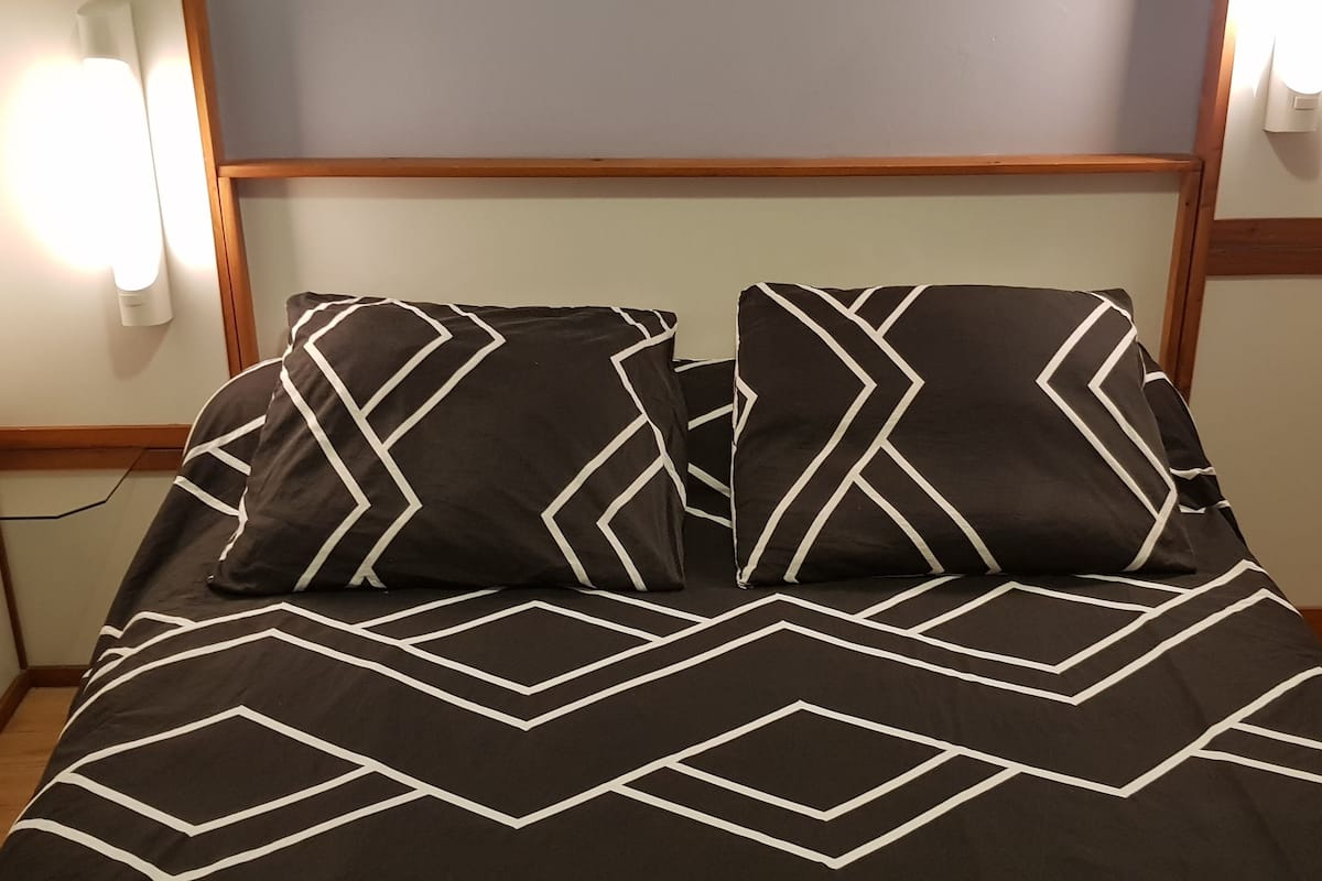 A bed with a geometric patterned bedspread and two matching pillows, a red stool, and wall-mounted light fixtures.