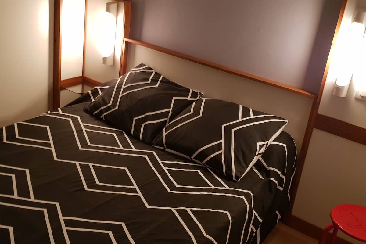 A bed with a geometric patterned bedspread, a wooden headboard, bedside lamps, and a small red stool.
