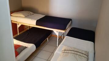 A small, compact room with two single beds, each with a pillow and a blanket. The room has a tiled floor and plain walls.