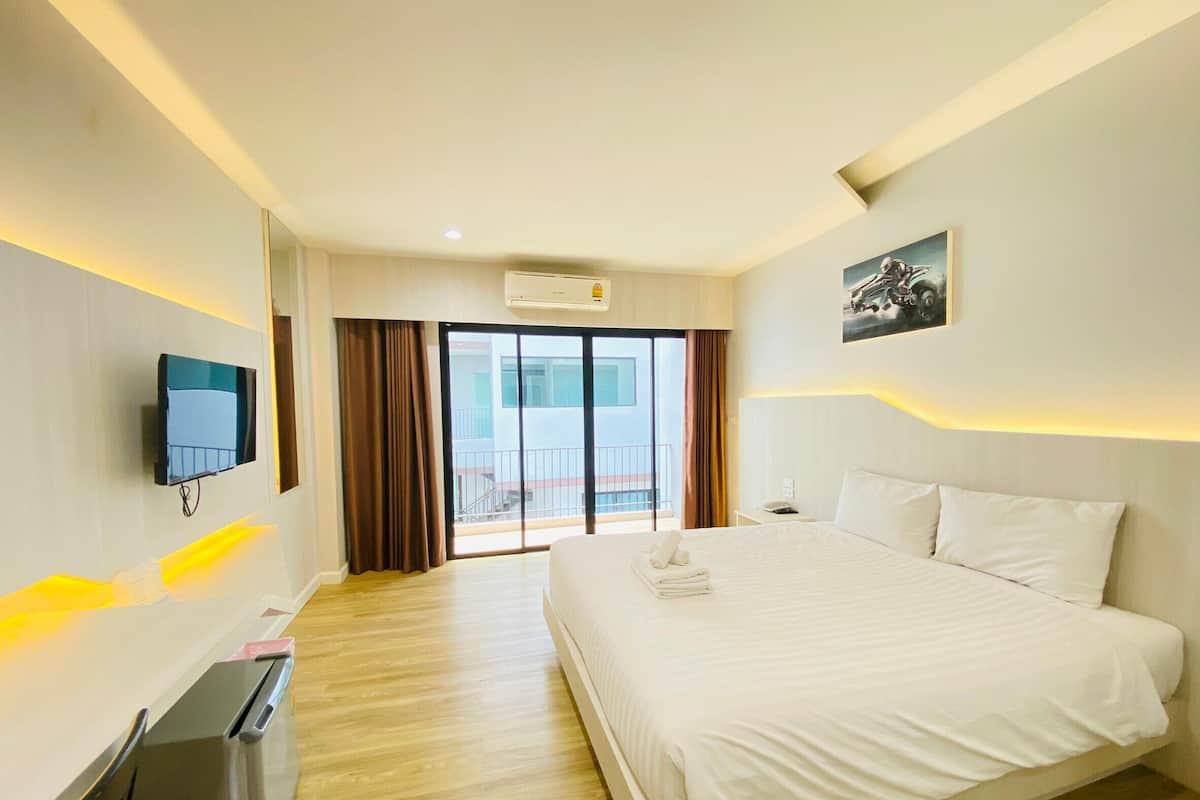 A hotel room with a large bed, a flat-screen TV mounted on the wall, a desk with a computer, and a sliding glass door leading to a balcony.