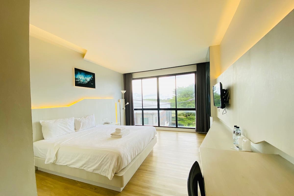 A modern hotel room with a large bed, a desk, a chair, a TV, and a large window with a view of the city.