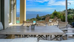 A covered outdoor seating area with a table and chairs, overlooking a pool and a scenic view of the sea and mountains. - Apeiron II Villa - Two Bedroom Villa, Sleeps 4 (Paxi)