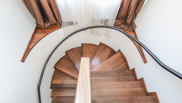Un escalier en colimaçon en bois, doté d’une rampe noire et bordé d’un mur blanc.