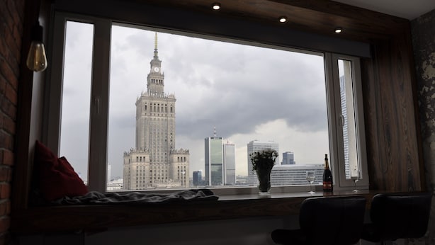 A room with a large window overlooking a cityscape, including a prominent building with a clock tower. - Panda Apartments Bagno- Centrum (Warsaw)