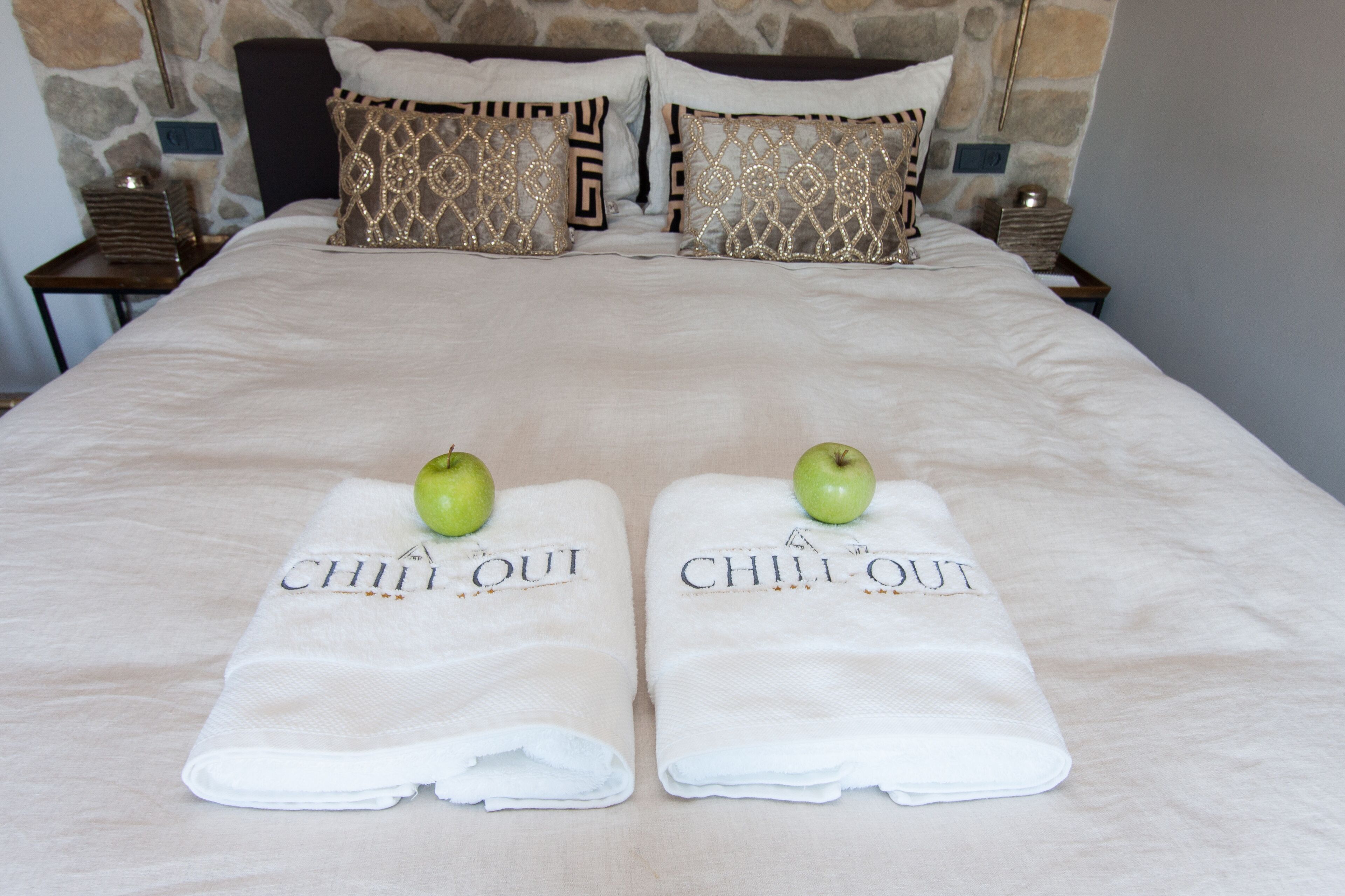 A neatly made bed with two towels folded on top, each with a green apple and the text "CHILI OUT" on them.