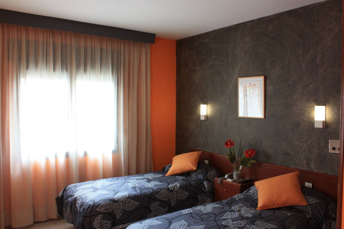 Ein Hotelzimmer mit zwei Betten, einem großen Fenster mit Vorhängen, einer Vase mit Blumen und einem gerahmten Bild an der Wand.