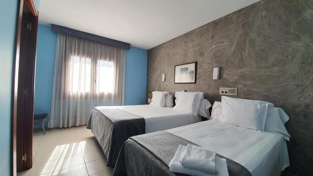 Une chambre d’hôtel avec deux lits, une grande fenêtre avec des rideaux, un tableau accroché au mur et une petite table surmontée d’un vase.
