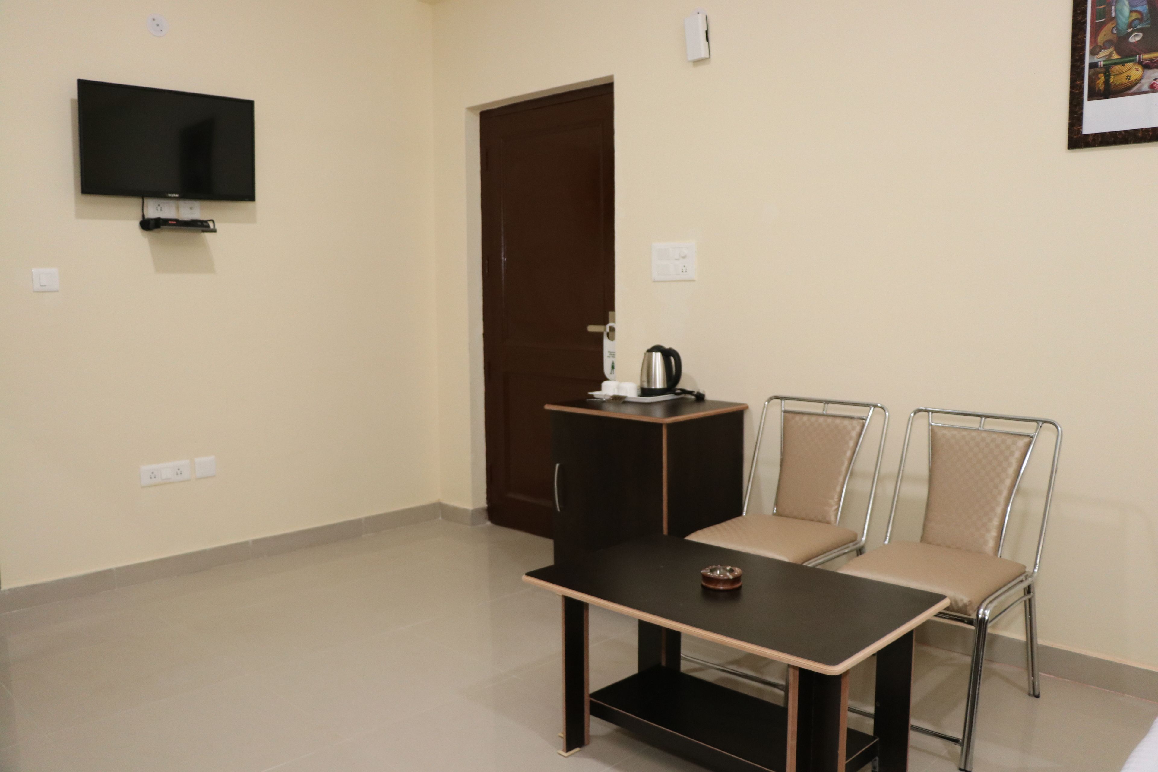 A room with a flat-screen TV mounted on the wall, a dark wooden table with a small tray, two metal-framed chairs with beige cushions, and a dark wooden door.